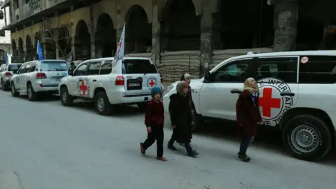 Getty Images Syrian children walk past vehicles of the UN and the International Committee of the Red Cross (ICRC) delivering humanitarian aid in the Syrian town of Douma