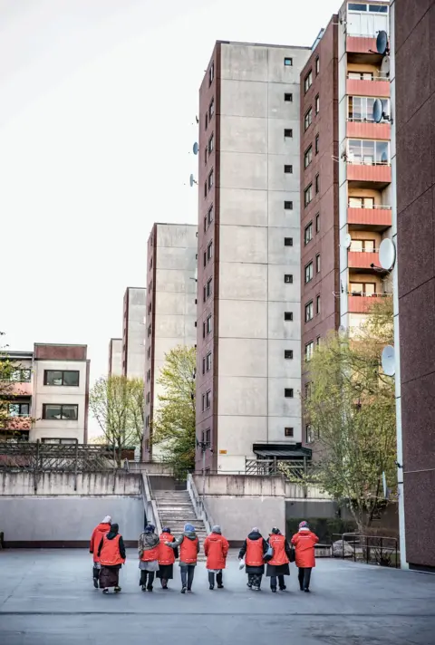 Anette Nantell A line of women in red coats make their way through a housing development.