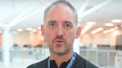 Northamptonshire Police Man with short hair and beard, wearing a lanyard stands in a large building