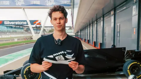 Alex Witty stands next to the Silverstone race track holding one of his new trainers with a Formula One car in the background