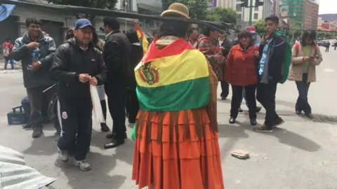 Elsa Andrade stands in the street, wearing a traditional skirt and draped in a Bolivian flag