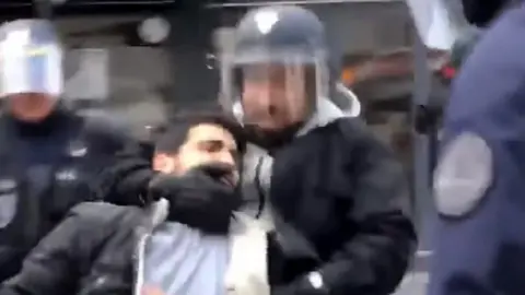 TAHAR BOUHAFS a pixelalted image shows a bearded man wearing a hoody and a riot police helmet grabbing a protester by the neck