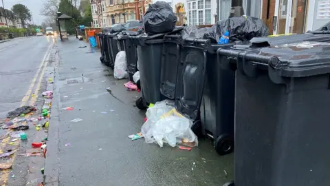 BBC A row of overflowing bins along a Swindon road with cars and homes, rubbish going into the road