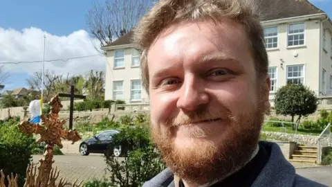 Rev Michael Darkins Rev Michael Darkins, a bearded white man, smiling at the camera. The background shows green bushes and a property with windows.