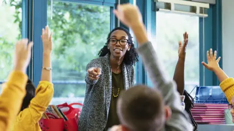 Getty Images A teacher wearing black glasses and a grey cardigan smiles as she points towards a student with their hand in the air, surrounded by classmates also with their hands up.