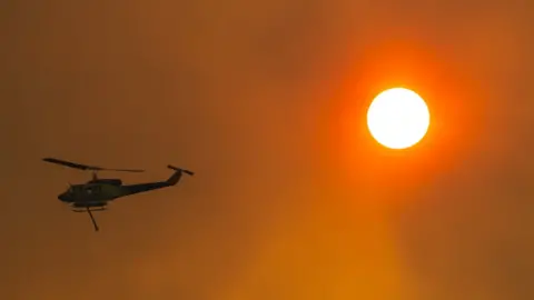 Getty Images A water-bombing helicopter silhouetted on a red, smoke-filled sky next to the sun