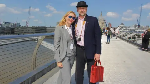 A man and woman stand on millennium bridge. Man is in a top hat and suit, woman is in a suit.