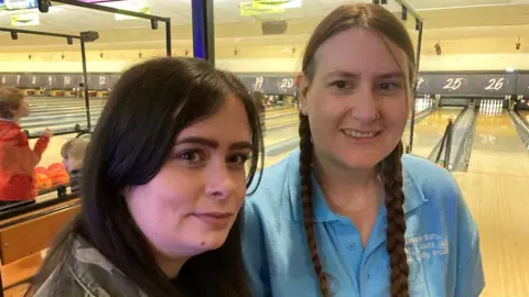 Bron Edwards and Becky Harrison standing together at the 10 pin bowling lane. Bron is the support worker for Becky Harrison who is registered blind