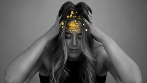 Sammy Weston  Black and white image of a woman with her hands on the side of her head. The only form of colour in the picture is the gold sequins on her forehead.