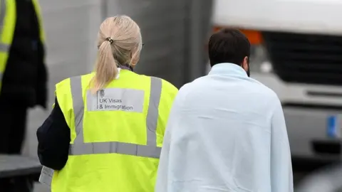 Getty Images A woman in a hi-vis UK Visas and Immigration jacket walks beside a man wrapped in a towel