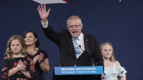 Brook Mitchell / Getty Scott Morrison waves to crowd from a podium with his wife and two daughters