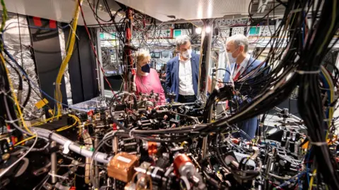 Getty Images Angela Merkel stands next to two men in front of a complex, somewhat messy array of pipes and wires