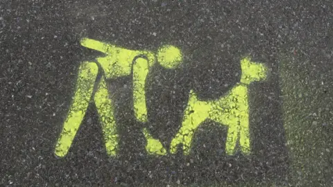 Stencil in yellow on tarmac of person picking up dog poo
