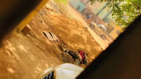 Reuters A soldier's body lies next to a mound of soil while a soldier uses a building for cover across the street in Khartoum on 15 April 2023, as fighting escalated