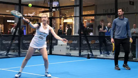 Getty Images Tia and Andy Murray. Tia, a woman, is holding a black padel bat with an outer green rim - in her right hand mid-shot. Her left hand is outstretched. Andy Murray, a man is standing to the right watching her hit the ball, he is wearing a grey long sleeved sports top and black tracksuits. They are both standing on a blue court. There are a few people in the background watching them play padel.