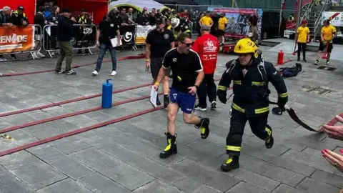 Ronnie Sherratt Man in firefighter uniform running whilst dragging a hose behind him, A man is running beside him.