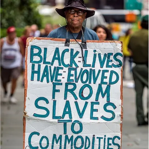 Getty Images Activist holding sign reading BLACK LIVES HAVE EVOLVED FROM SLAVES TO COMMODITIES