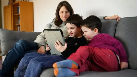 Getty Images Two young children and their mother look at a tablet computer on a sofa