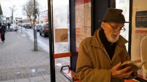 Getty Images Elderly man wearing a hat and glasses reads a book at a bus stop