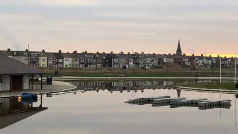 Duncan Leatherdale A boating pond with houses beyond reflected on the water