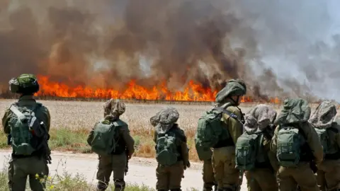 Getty Images Israeli soldiers walk amidst smoke from a fire in a wheat field near the Kibbutz of Nahal Oz, along the border with the Gaza Strip, on May 14, 2018