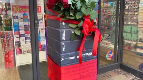 STEVE MARTIN/ BBC  Tributes laid by the card shop in which Mr Quiqley died. Roses and red ribbon are attached to the bollard.