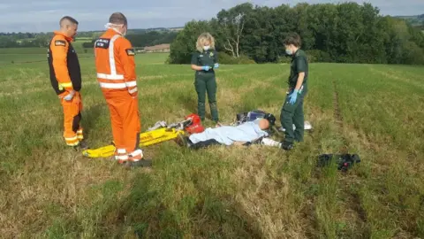 Northants Hunt Sabs Air ambulance personnel, two in orange hi-viz and two in green paramedic clothing attend to a man lying on the floor under a blue blanket