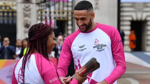 Getty Images The first Baton-bearer, double Paralympic gold medallist Kadeena Cox (L) hands over the baton to Team England squash player Declan James as Britain's Queen Elizabeth II launches the Queen's Baton Relay for the Birmingham 2022 Commonwealth Games, from the forecourt of Buckingham Palace in London on October 7, 2021