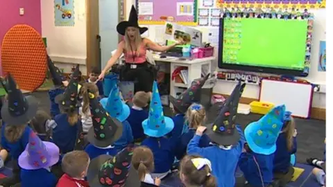 A class of children wearing witches hats listening to their teacher read a story