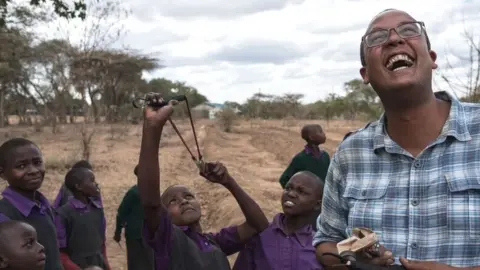 BBC A Kenyan man laughs as children aim a catapult at the sky