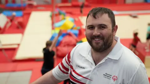 John Fairhall/BBC Alex Row, head coach in his coaching kit. He is standing in front of gymnastics matting and equipment.