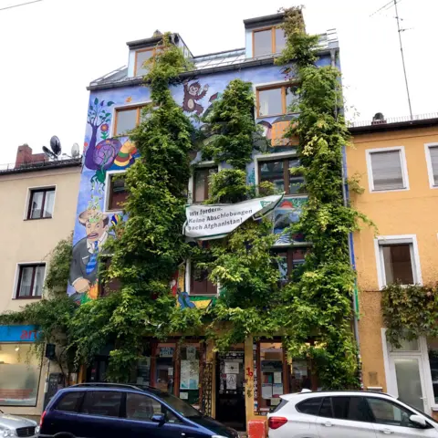 Ligsalz8 is pictured covered in green climbing plants with brightly-painted murals, in stark contrast to the standard terraced buildings on either side