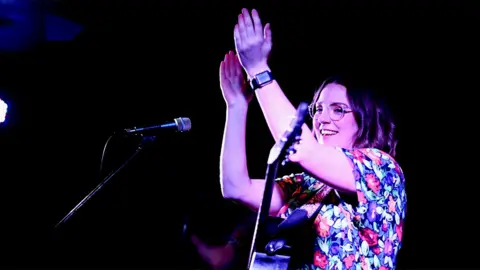 Symphorien Demores Lucy is wearing a floral top, with a guitar around her, clapping as she sings into the two microphones in front of her. She is wearing a watch on her left wrist. The background is plain black.