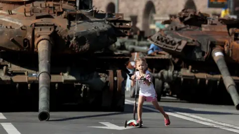 CATHAL MCNAUGHTON Girl on scooter in front of armoured tanks