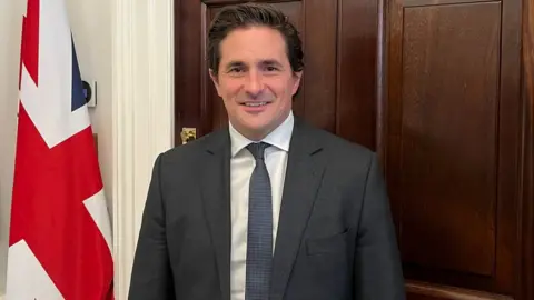 BBC Johnny Mercer, with brown hair wearing a grey suit with a white shirt and a navy tie with tiny white spots
