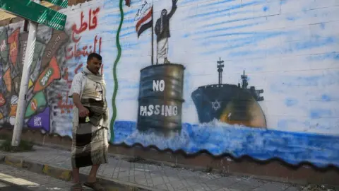 EPA A Yemeni man walks past a mural showing a Houthi fighter stopping an Israeli ship in the Red Sea, in Sanaa, Yemen (19 February 2024)