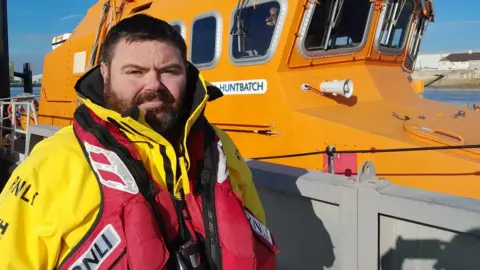 RNLI/Tom Collins Robbie Maiden stands next to a lifeboat