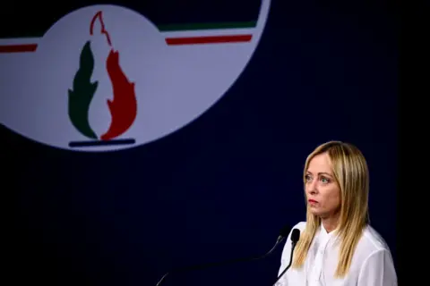 Getty Images Giorgia Meloni standing next to a large sign of her party's logo, the tricolour flame