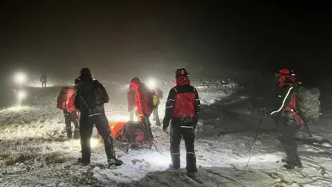 Keswick Mountain Rescue Volunteers in red and black coats and lit by headtorches push a stretcher across snow and rocks