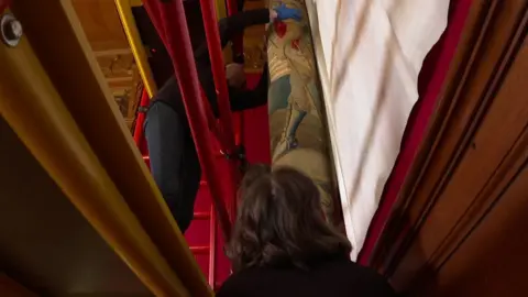 Woman on scaffolding prising tapestry off wall