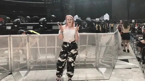 Beth Beth is posing for a photo at a K-Pop BTS concert. She has a pink bob and is posing with her fingers held up in two v shapes. She's wearing a white top and black and white cargo pants. She is stood in front of a metal gate that is shoulder height. In the background you can see all the concert stage, stewards and lights.