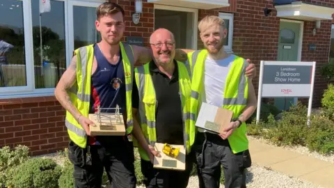 BBC Jack Aldis, Kevin Chapman and Jamie Powell stand with their arms around one another holding their construction style trophies