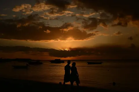 Getty Images Two people walk along a beach at sunset. Their silhouettes are seen against the orange, setting sun.