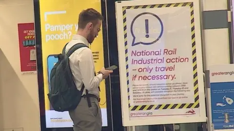 BBC man checks phone in front of information sign about rail strike