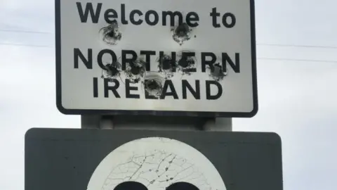 BBC bullet holes riddle the Welcome to NI sign