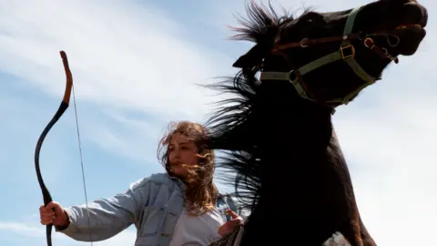 Bertan Berber/ bb.stunts Catriona Scott, a 30-year-old white woman with long brown hair, rides a black horse as it rears up. She holds a bow in her right hand, looking to the side. Catriona wears her hair loose and it's caught in the wind along with the horse's mane. She wears a denim jacket, jeans and a white top. She's pictured outside on a sunny day against a blue sky dusted with clouds.