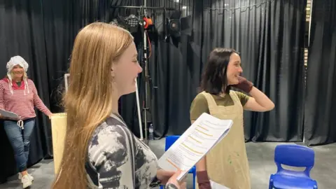 Three actors, one holding a script, are rehearsing with black curtains and spotlights in the background