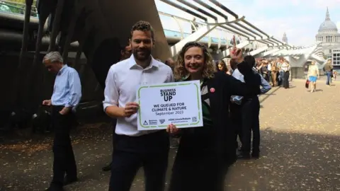 A woman and man hold a sign in front of millennium bridge, saying 'businesses stand up'. She is pointing up. A queue of demonstrators is seen behind them.