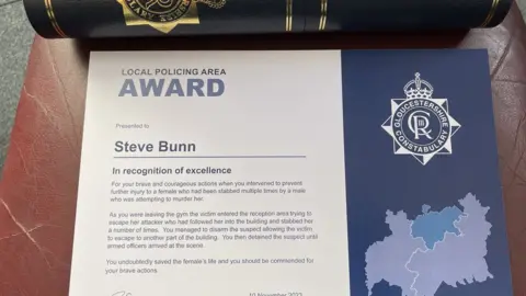 Steve Bunn A certificate for Steve Bunn from the Gloucestershire Constabulary "in recognition of excellence", written in blue on a white background, with a smart black and gold tube in the background to contain it.