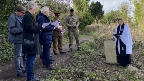 BBC A clergyman reads from a Bible over a grave as uniformed soldiers and a group of people look on